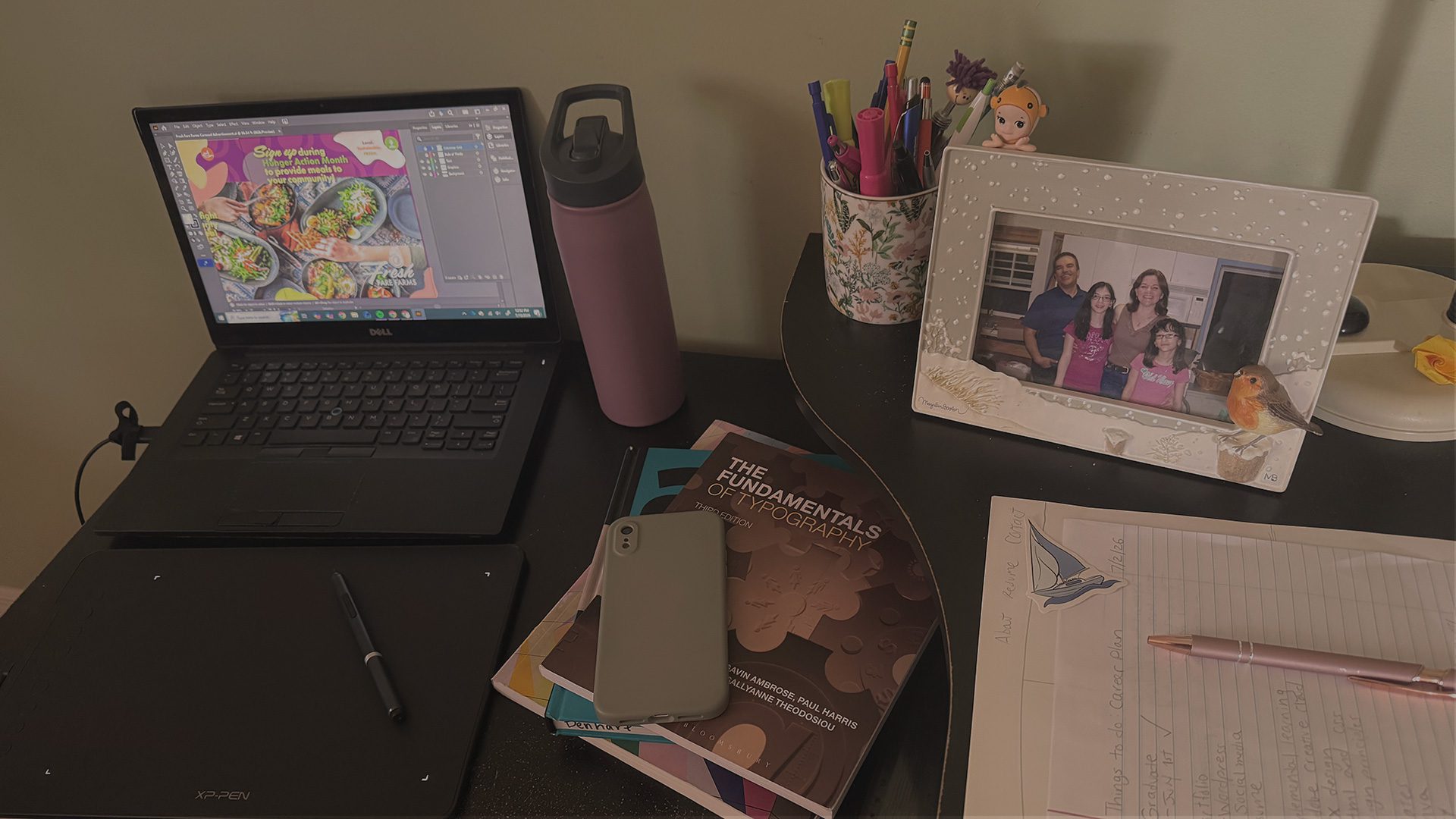 Graphic designer desk with adobe illustator open on a Dell laptop, a drawing tablet, design books, and a water bottle. A flowery mug filled with writing tools, an image of a family, and neatly stacked papers with a pen on top are to the right of the image.