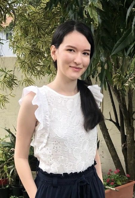 Picture of Daniella Cicirello posing with her hands in her trousers in a garden. She has black hair in a ponytail, pale skin, and is wearing a white floral, short-sleeve shirt tucked into navy trousers.